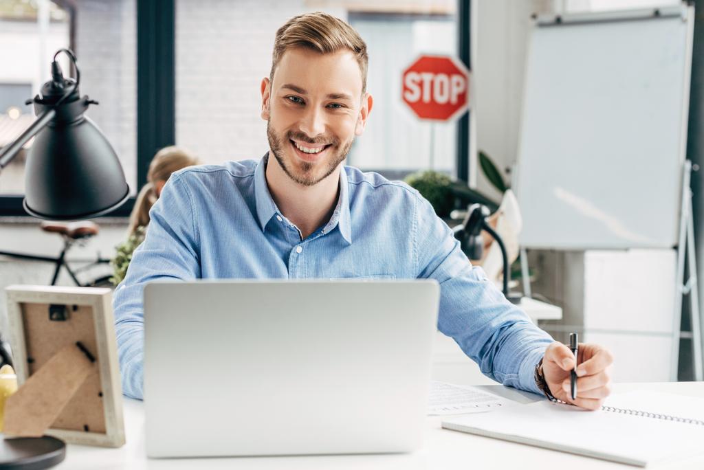 όμορφος χαμογελαστός νεαρός επιχειρηματίας χρησιμοποιώντας φορητό υπολογιστή και λήψη σημειώσεων στο χώρο εργασίας  - Φωτογραφία, εικόνα