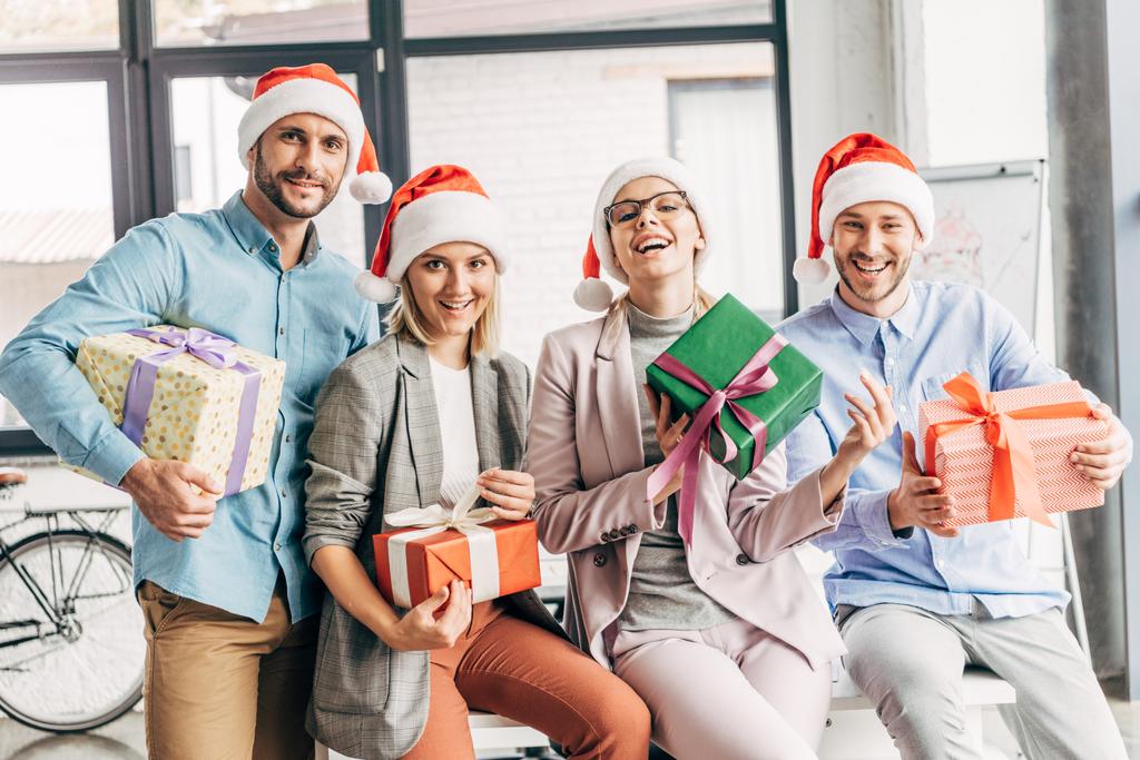 onnellisia työtovereita jouluhatuissa pitäen lahjoja ja hymyillen kameran edessä toimistossa
 - Valokuva, kuva