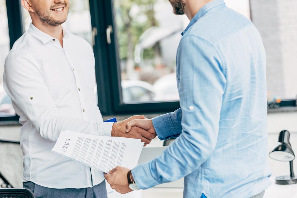 обрезанный снимок улыбающихся бизнесменов, держащих контракт и пожимающих руки в офисе
 - Фото, изображение