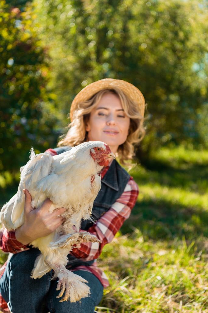 Lächelnde Frau mit Strohhut und weißem Huhn im Freien  - Foto, Bild