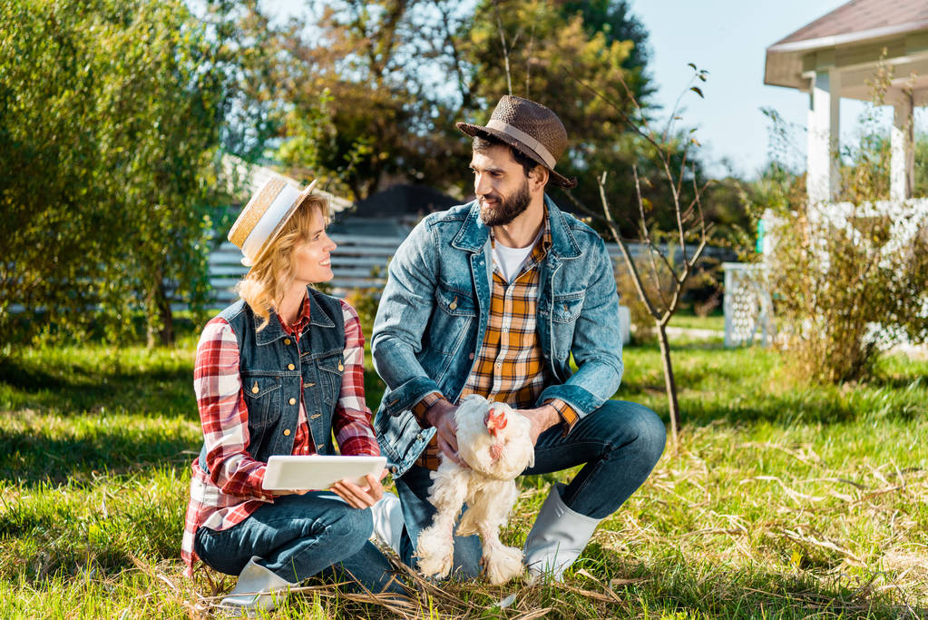 Lächelnder Landwirt hält Hühner, während seine Freundin auf dem Hof ein digitales Tablet benutzt - Foto, Bild