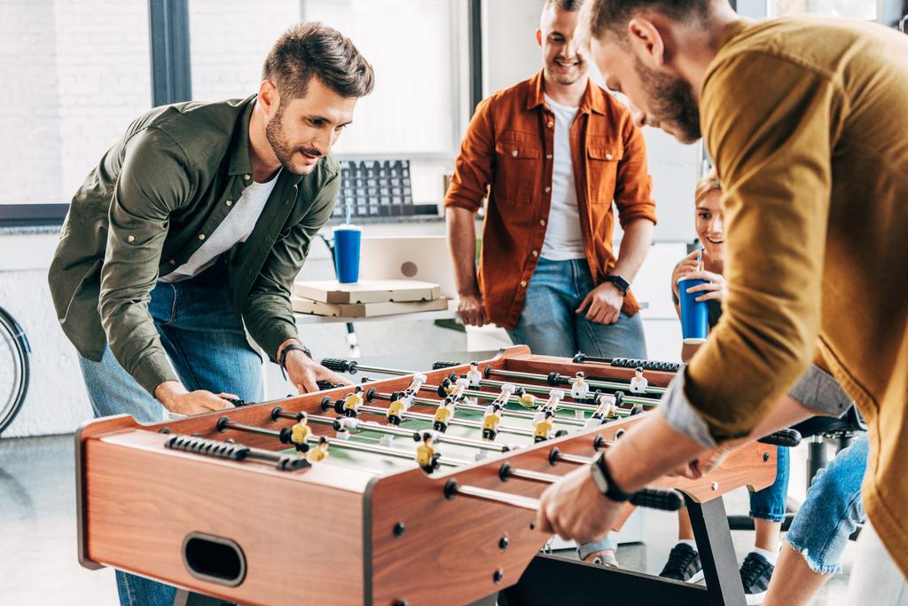 группа молодых случайных бизнесменов, играющих в настольный футбол в офисе и веселящихся вместе
 - Фото, изображение