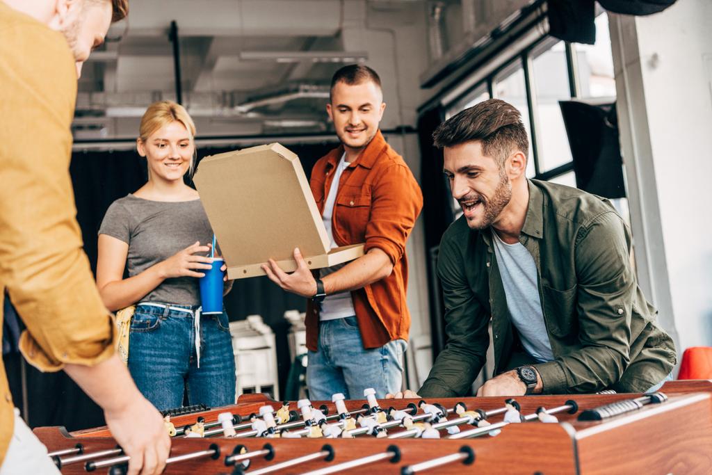 grupo de jóvenes empresarios casuales jugando al futbolín con pizza en la oficina y divirtiéndose juntos
 - Foto, imagen
