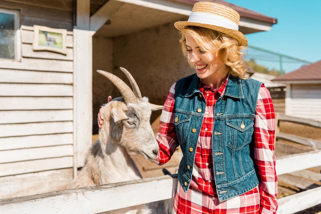 mooie lachende vrouw aanraken van geit in de buurt van houten hek op boerderij - Foto, afbeelding