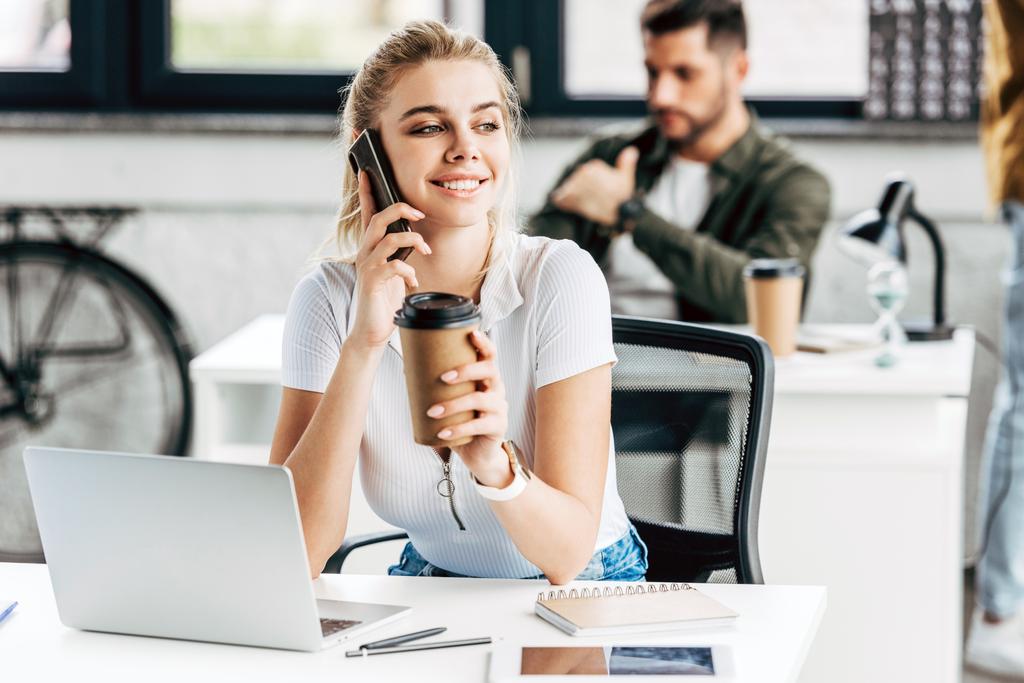 aantrekkelijke jonge vrouw praten via de telefoon en het drinken van koffie op kantoor met collega op achtergrond - Foto, afbeelding
