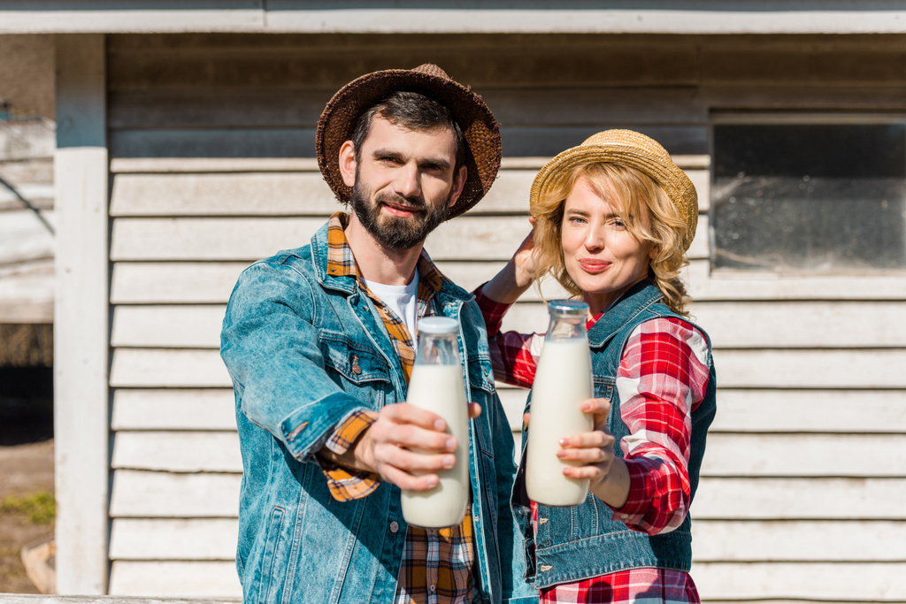 ενηλίκων ζευγάρι των αγροτών στην άχυρο καπέλα ψάθινο καπέλο δείχνει μπουκάλια γάλακτος στο αγρόκτημα - Φωτογραφία, εικόνα