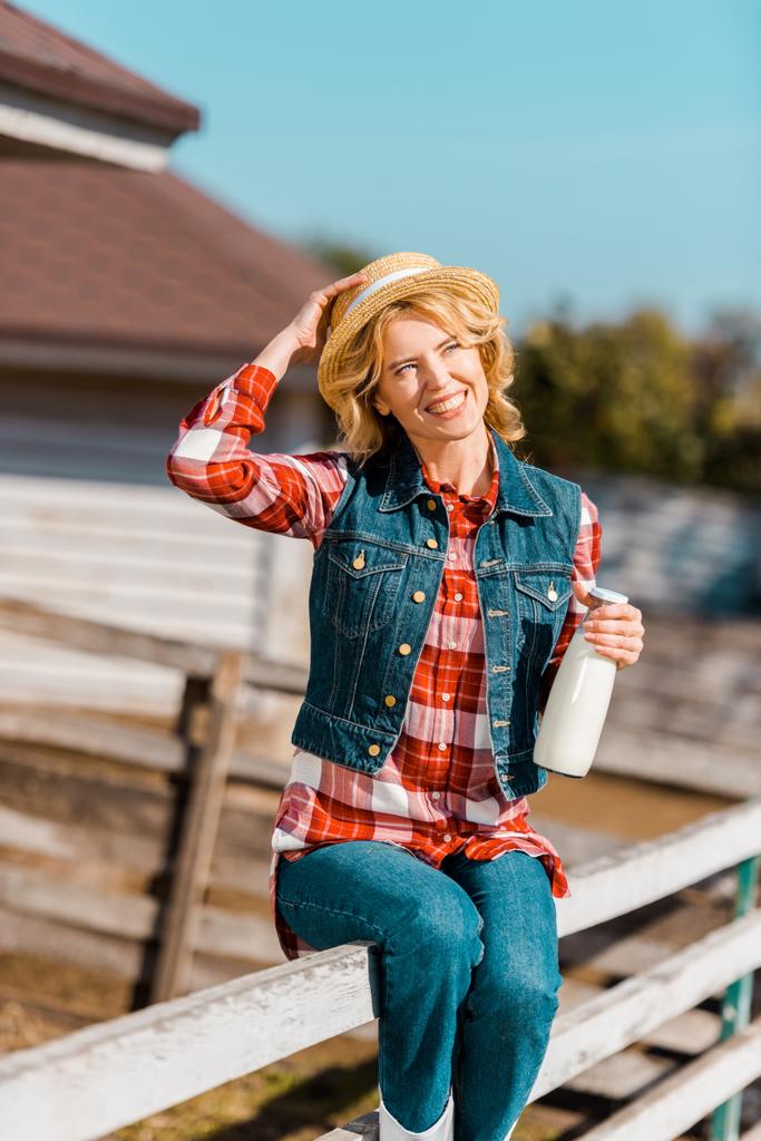 happy female farmer holding bottle of milk and sitting on wooden fence at ranch - Photo, Image