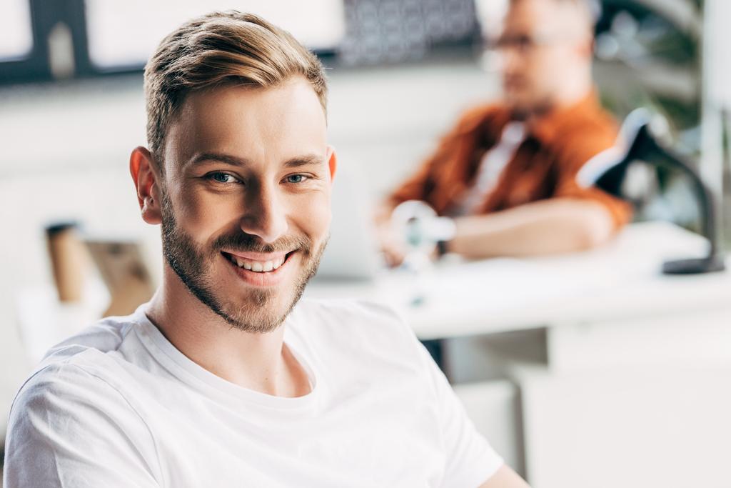 handsome young businessman looking at camera with colleague sitting blurred on background at office - Photo, Image