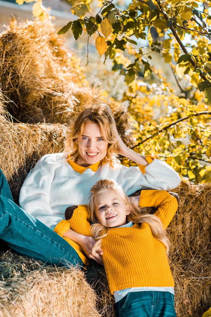 fröhliche Frau posiert mit kleiner Tochter auf Heuhaufen auf Bauernhof - Foto, Bild