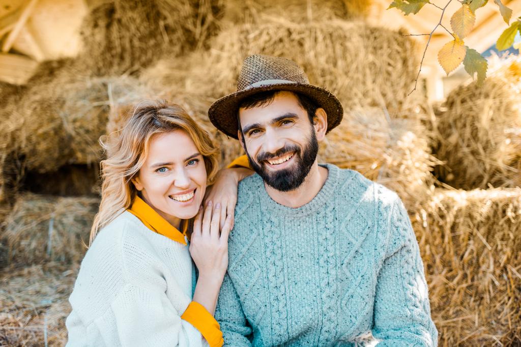 portrait of happy couple of farmers looking at camera while sitting on stacks of hay at farm  - Photo, Image