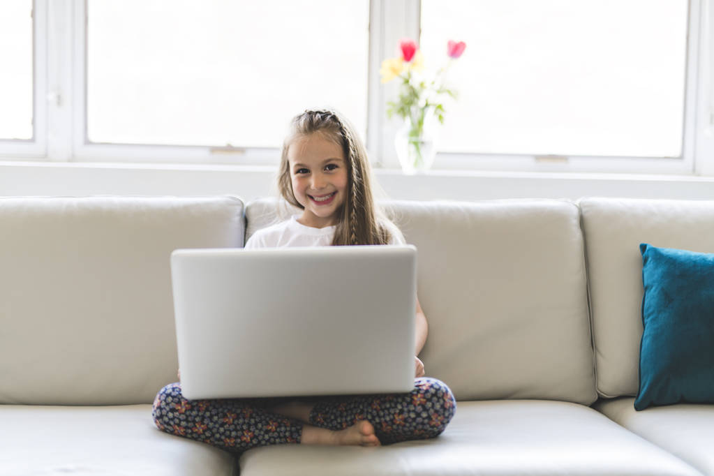 Χαρούμενος στο σπίτι το μικρό κορίτσι που εργάζονται με ένα laptop στον καναπέ - Φωτογραφία, εικόνα