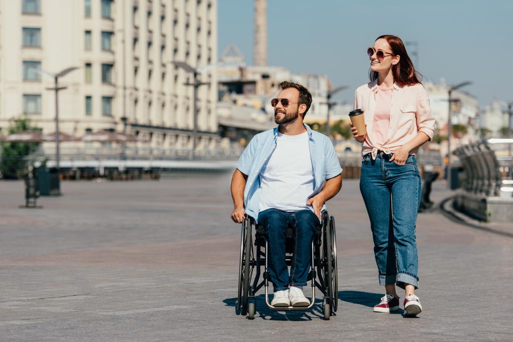 guapo novio en silla de ruedas y novia con café en taza de papel teniendo caminar en la calle y mirando hacia otro lado
 - Foto, Imagen