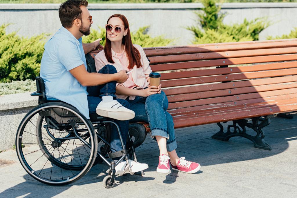 handsome boyfriend in wheelchair with smartphone and girlfriend with coffee to go spending time on street - Photo, Image