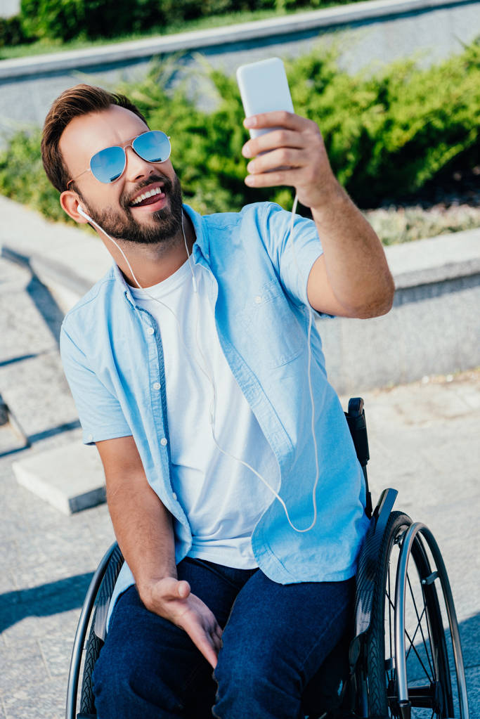 pohledný muž v invalidní vozík držet jazyk ven a při zohlednění selfie s smartphone na ulici - Fotografie, Obrázek