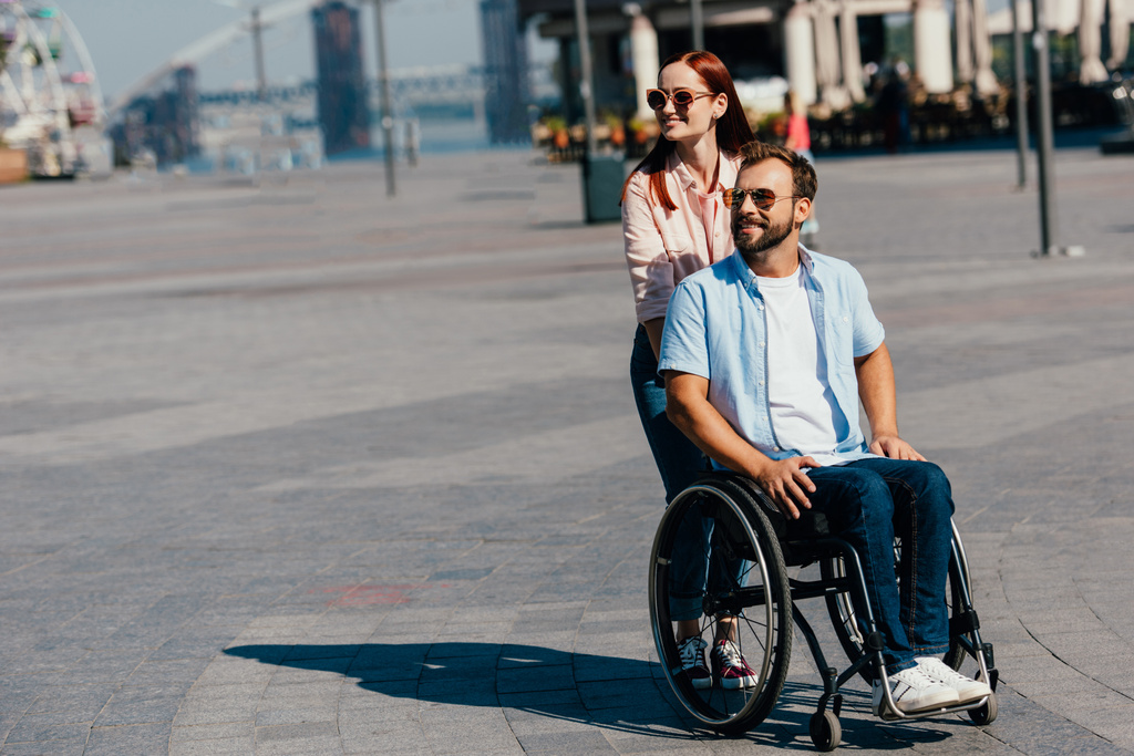 attractive girlfriend in sunglasses pushing disabled handsome boyfriend in wheelchair on street - Photo, Image