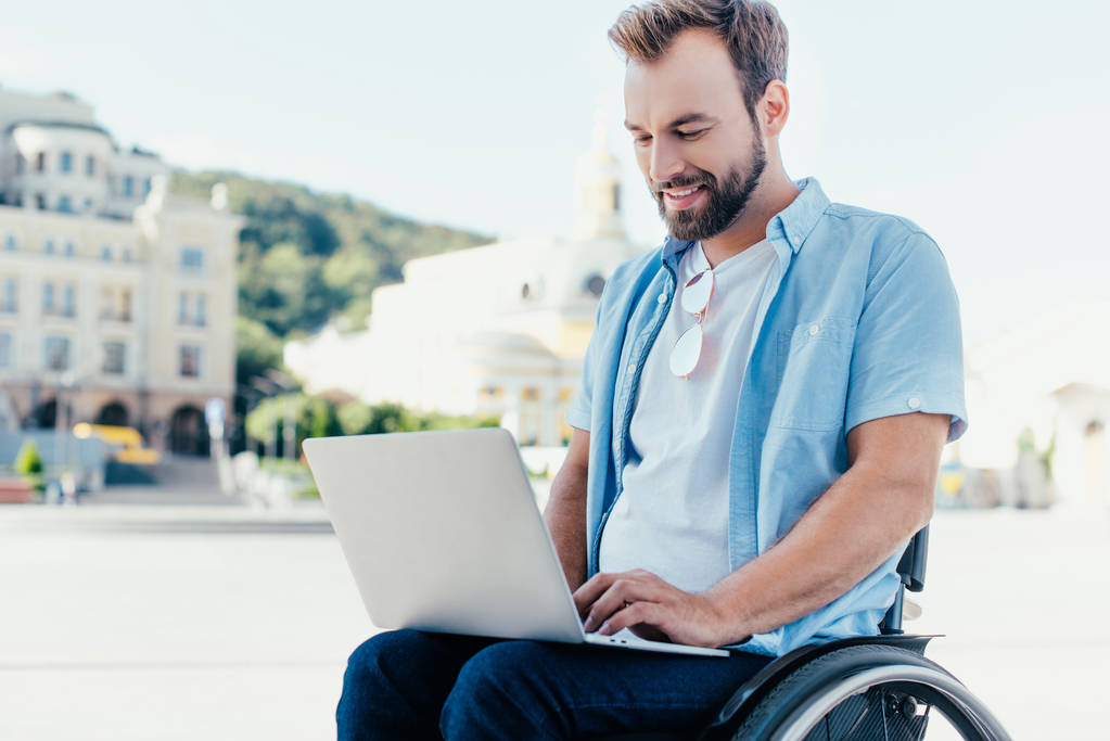 vrolijke knappe man in rolstoel met behulp van laptop op straat - Foto, afbeelding