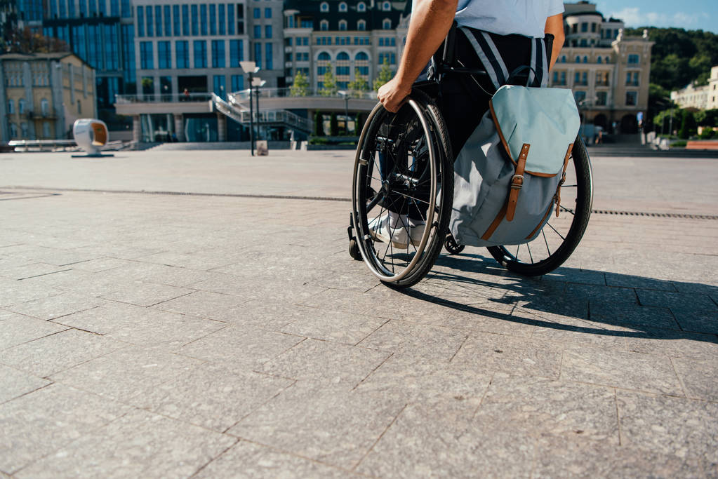 περικομμένη εικόνα του ανθρώπου που χρησιμοποιούν αναπηρικό αμαξίδιο με την τσάντα στην πόλη - Φωτογραφία, εικόνα