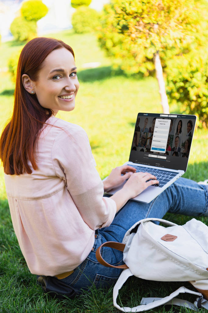 uśmiechający się atrakcyjną kobietę za pomocą laptopa z linkedin ładowane strony park i patrząc na kamery - Zdjęcie, obraz