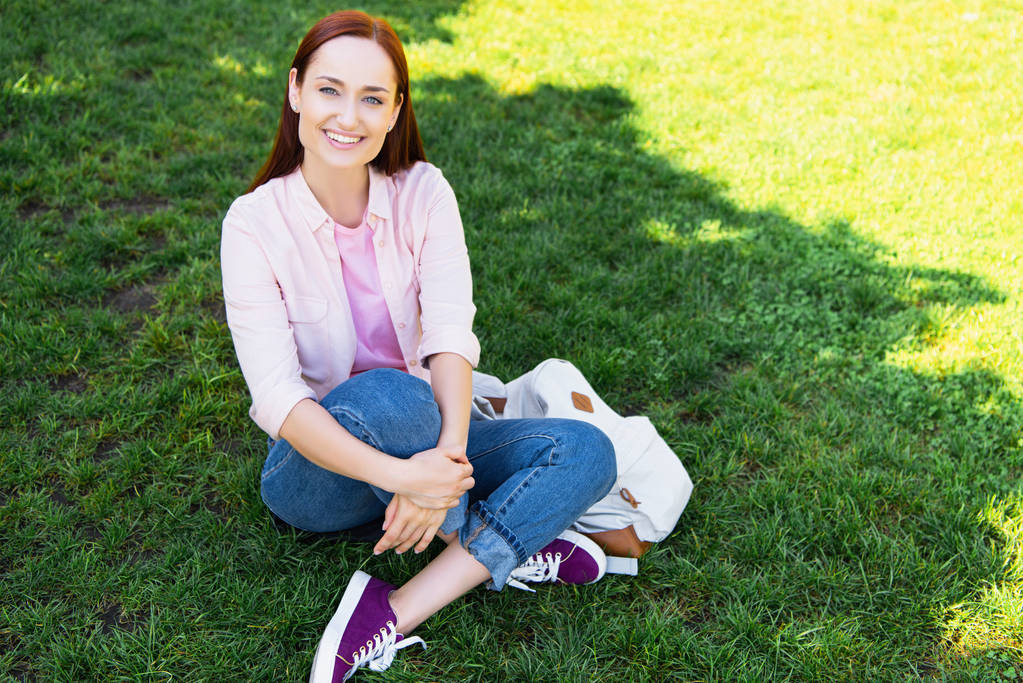 uśmiechający się atrakcyjna kobieta siedzi na zielonej trawie w parku i patrząc na kamery - Zdjęcie, obraz