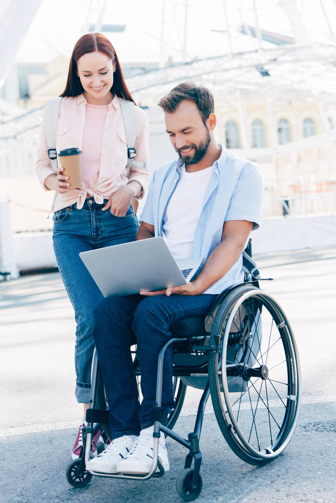 knappe vriendje in rolstoel met laptop en vriendin permanent met koffie te gaan op straat - Foto, afbeelding