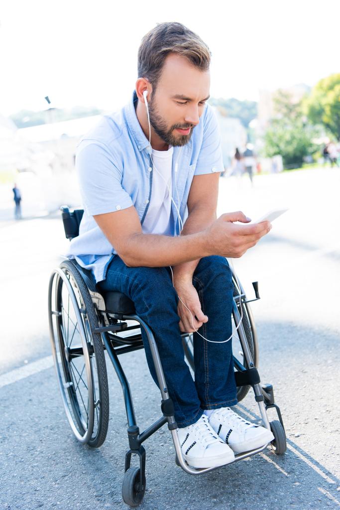 przystojny człowiek na wózku inwalidzkim, słuchanie muzyki ze smartfona na ulicy - Zdjęcie, obraz