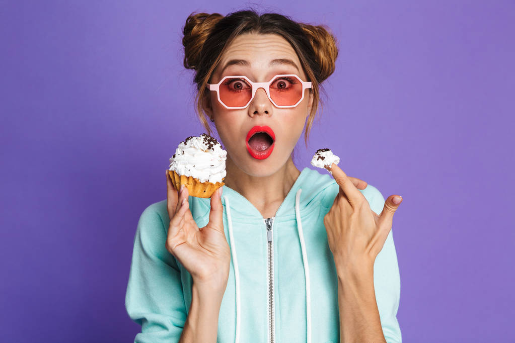 Retrato de una joven excitada con maquillaje brillante aislado sobre fondo violeta, sosteniendo cupcake
 - Foto, Imagen