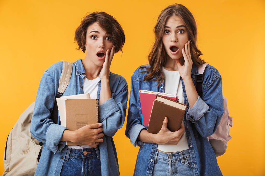 Зображення шокованих молодих жінок друзів ізольовано на жовтому фоні стіни, що тримає книги, які виглядають камерою
. - Фото, зображення