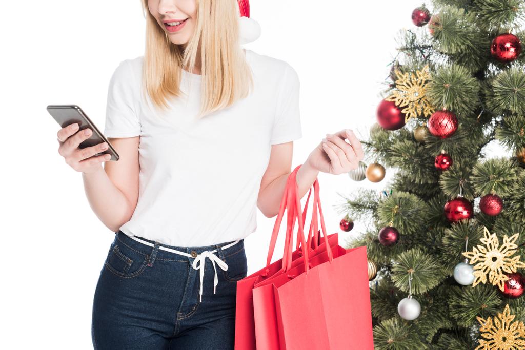 recortado tiro de mujer en sombrero de santa claus con bolsas de compras utilizando teléfono inteligente cerca de árbol de Navidad aislado en blanco
 - Foto, Imagen