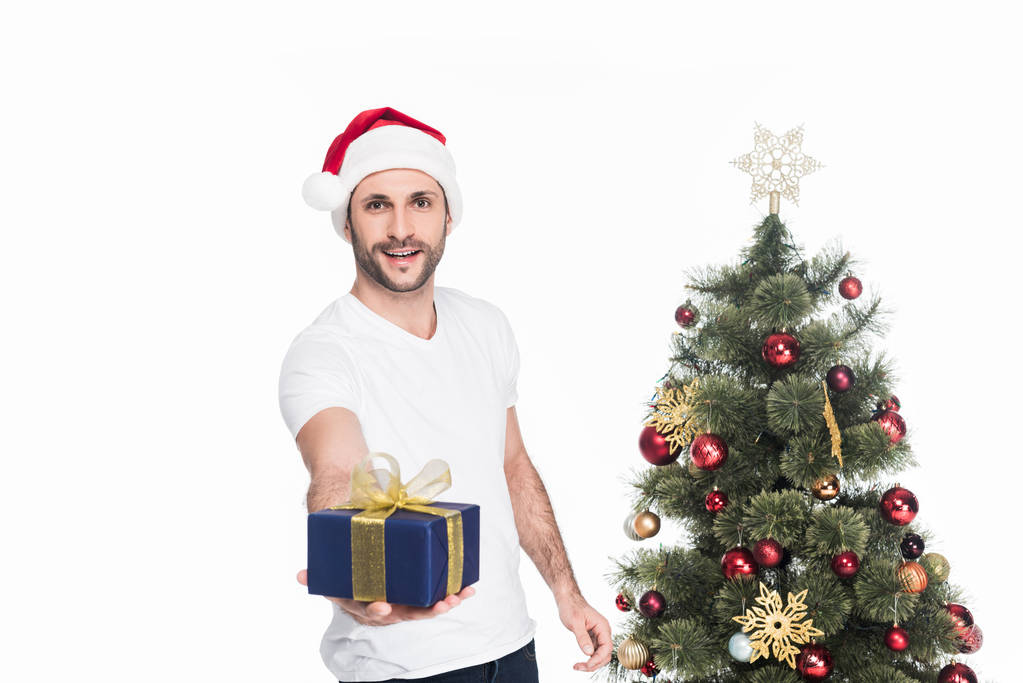 portrait of young man in samta claus hat with wrapped present standing at christmas tree isolated on white - Photo, Image