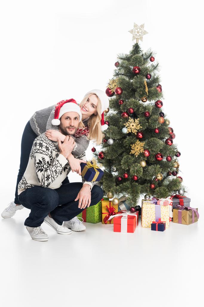 Paar in Pullovern und Weihnachtsmannmützen umarmen sich in der Nähe des Weihnachtsbaums - Foto, Bild