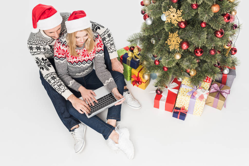Εναέρια άποψη του ζευγάρι σε καπέλα Άγιου Βασίλη χρησιμοποιώντας φορητό υπολογιστή κοντά στο χριστουγεννιάτικο δέντρο που απομονώνονται σε λευκό - Φωτογραφία, εικόνα