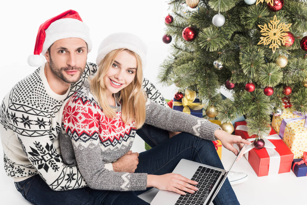 νεαρό ζευγάρι στο Βασίλη καπέλα με φορητό υπολογιστή κοντά στο χριστουγεννιάτικο δέντρο που απομονώνονται σε λευκό - Φωτογραφία, εικόνα
