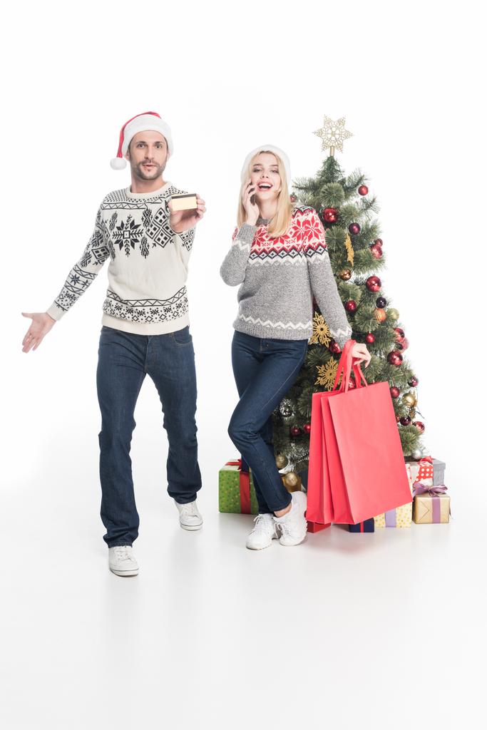 pareja en suéteres y sombreros de santa claus con bolsas de compras cerca del árbol de Navidad aislado en blanco
 - Foto, Imagen
