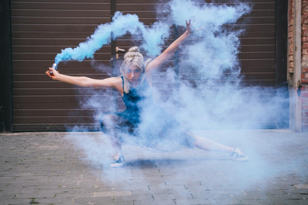 young contemporary dancer looking at camera and dancing in blue smoke on street  - Photo, Image
