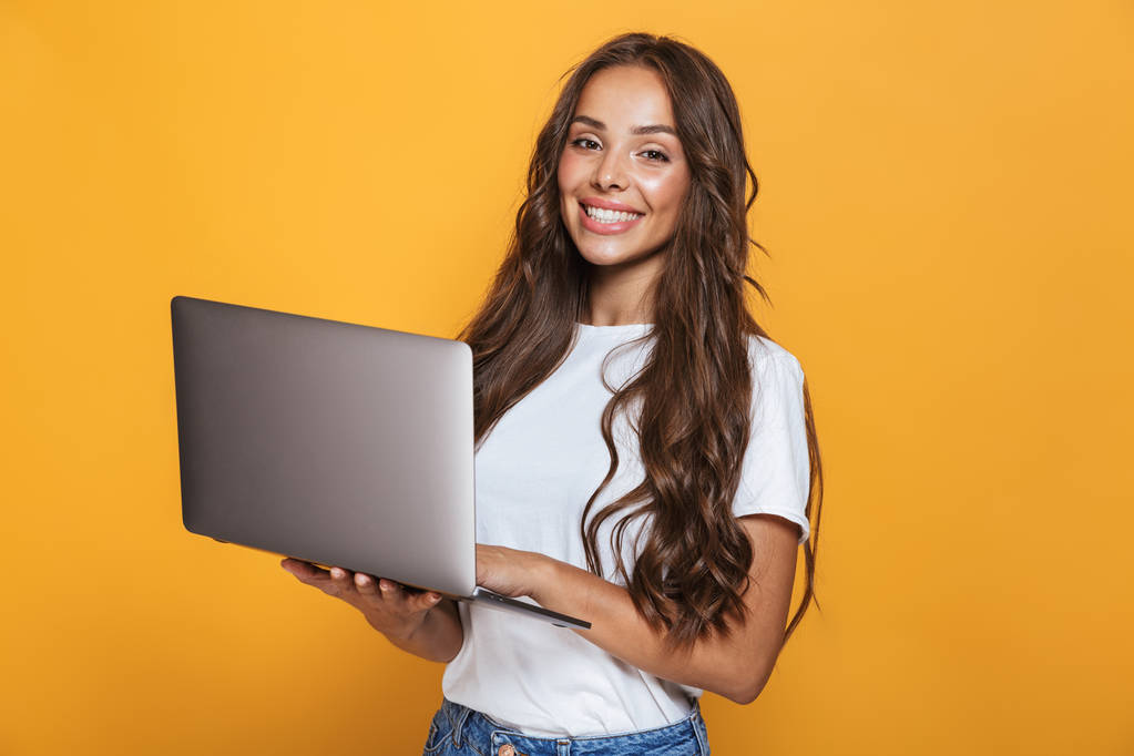 Retrato de mulher bonita 20s com cabelos longos sorrindo e segurando laptop cinza isolado sobre fundo amarelo
 - Foto, Imagem
