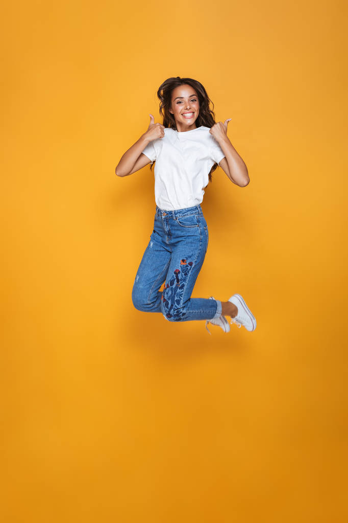Ganzkörperporträt eines fröhlichen Mädchens mit langen dunklen Haaren, das über gelben Hintergrund springt und Daumen nach oben zeigt - Foto, Bild