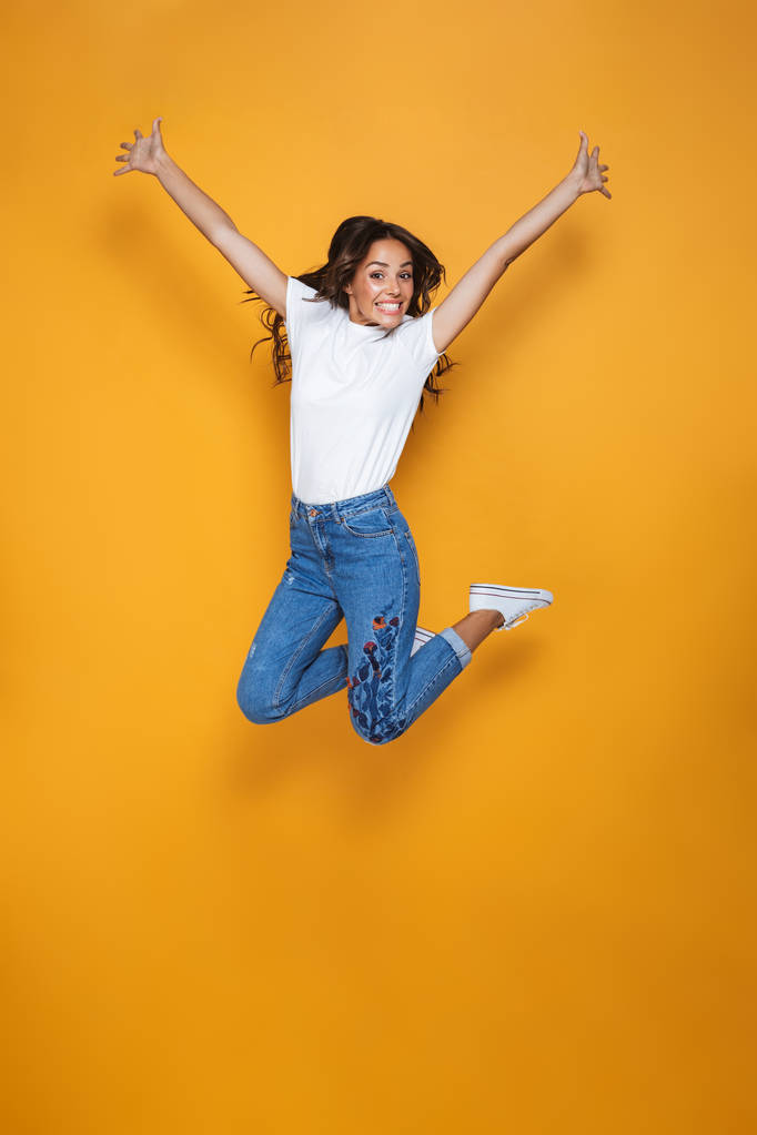 Retrato de larga duración de una chica alegre con el pelo largo y oscuro saltando sobre el fondo amarillo, celebrando el éxito
 - Foto, imagen
