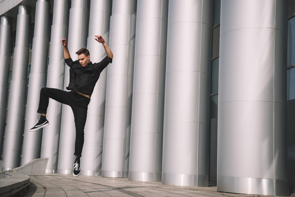 Красивый молодой человек в черной одежде танцует и прыгает возле колонн
   - Фото, изображение