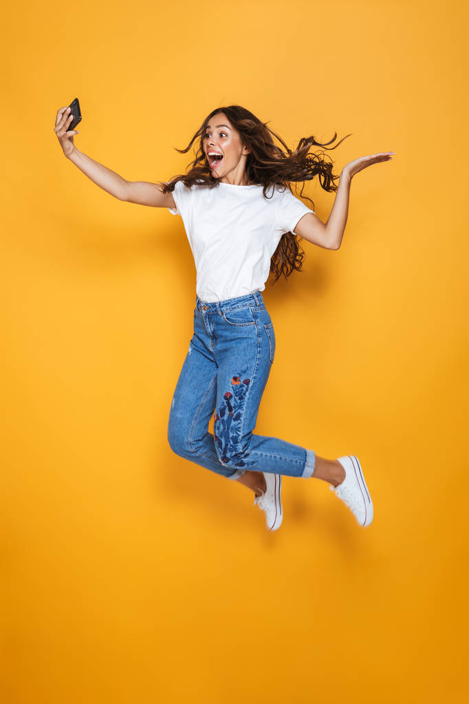 Full length portrait of a pretty girl with long dark hair jumping over yellow background, taking a selfie - Photo, Image