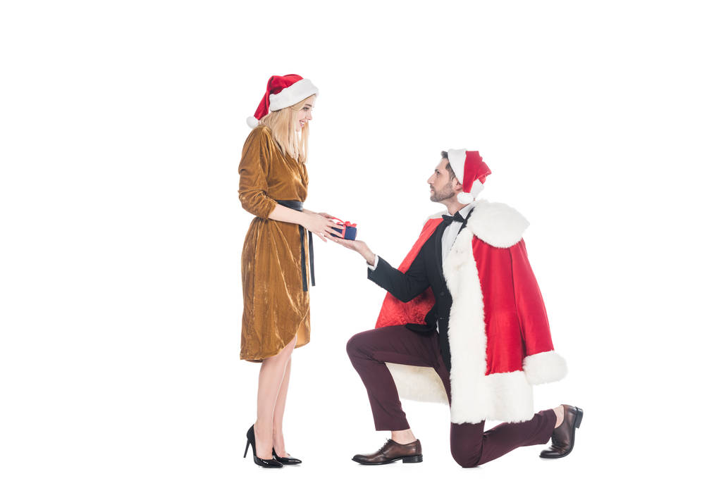 Mann im Weihnachtsmann-Kostüm überreicht Geschenk an Freundin mit Weihnachtsmann-Hut auf weißem Hintergrund - Foto, Bild