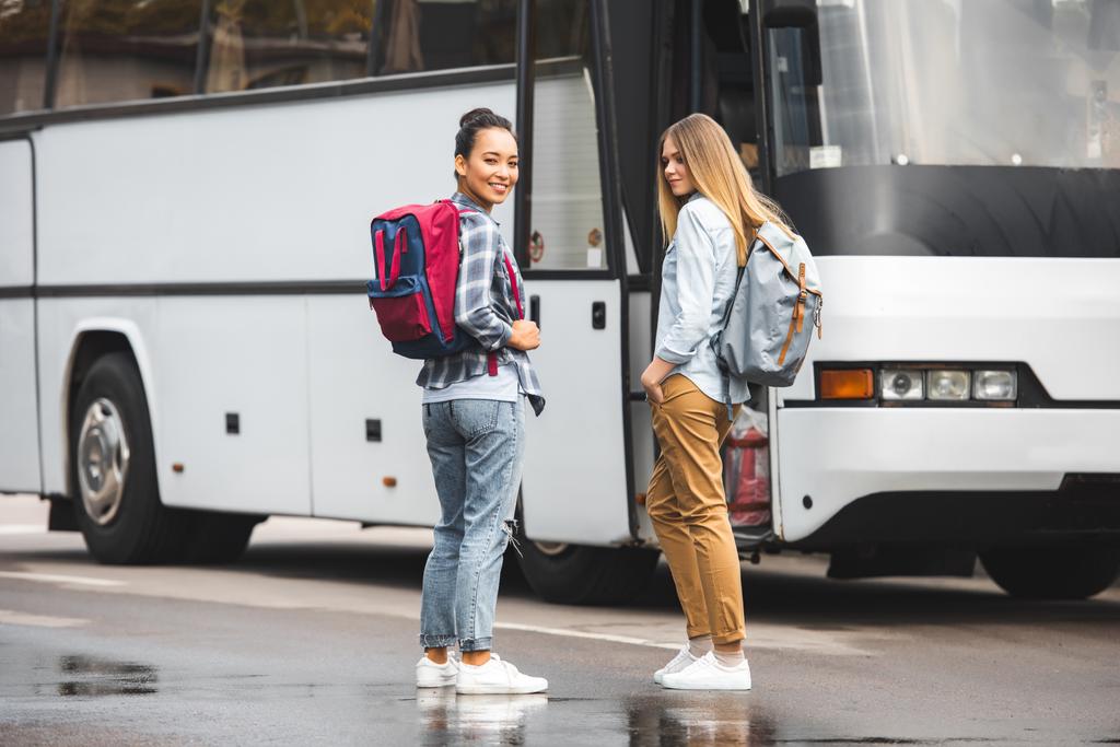 молодые мультиэтнические женщины с рюкзаками позируют возле туристического автобуса на городской улице
 - Фото, изображение