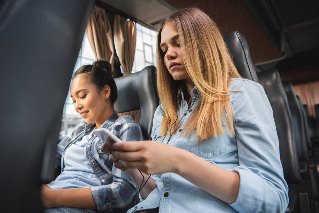 приваблива жінка сидить зі смартфоном, а її азіатська подруга сидить поруч під час подорожі на автобусі
 - Фото, зображення