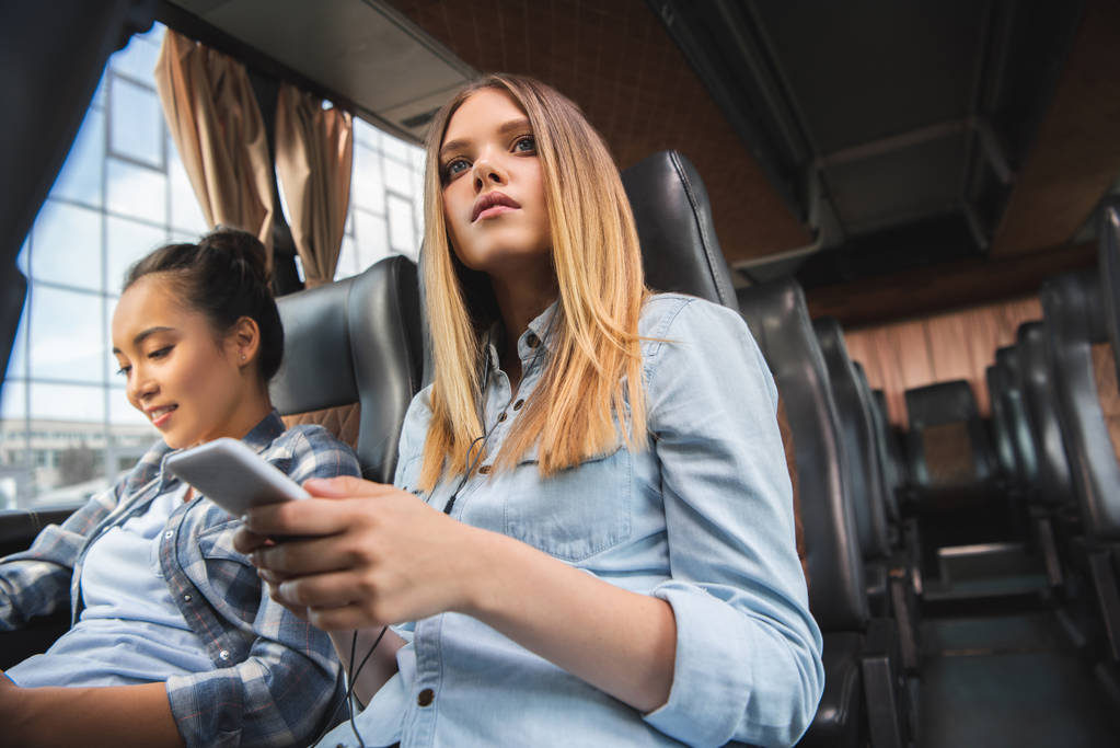 selektiver Fokus der Frau mit Smartphone, während ihre asiatische Freundin während der Fahrt im Reisebus in der Nähe sitzt - Foto, Bild
