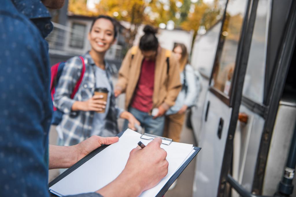 imagem cortada do controlador de ônibus de viagem escrevendo na área de transferência, enquanto os turistas de pé perto de ônibus na rua da cidade
  - Foto, Imagem
