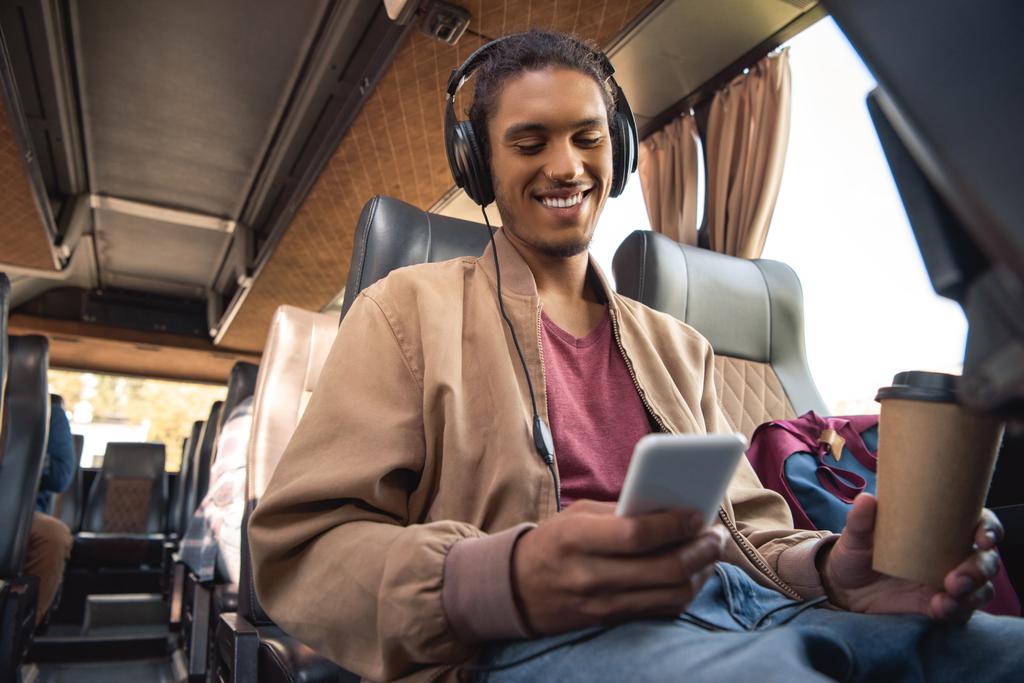 紙のコーヒー カップと旅行バスでスマート フォンに座っているヘッドフォンで笑みを浮かべて混血男 - 写真・画像