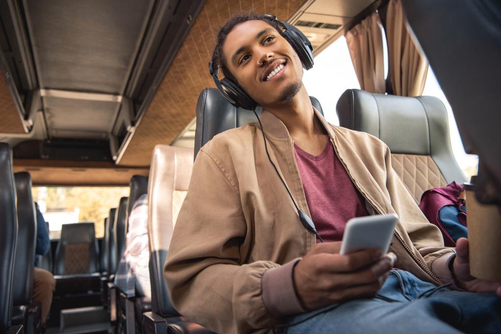 χαρούμενα μικτής φυλής ανθρώπου ακουστικά κάθεται με καφέ χαρτί Κύπελλο και smartphone σε λεωφορείο ταξιδιωτικό - Φωτογραφία, εικόνα