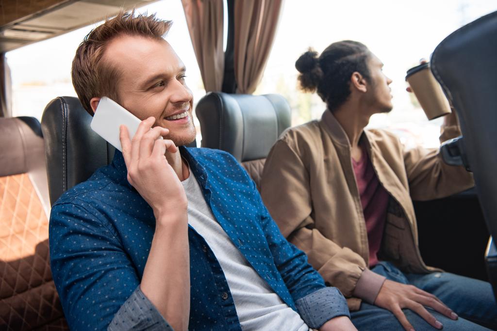 Χαμογελαστός άνθρωπος μιλώντας στο smartphone, ενώ ο φίλος πίνει καφέ κοντά κατά τη διάρκεια ταξιδιού σε λεωφορείο ταξιδιωτικό - Φωτογραφία, εικόνα