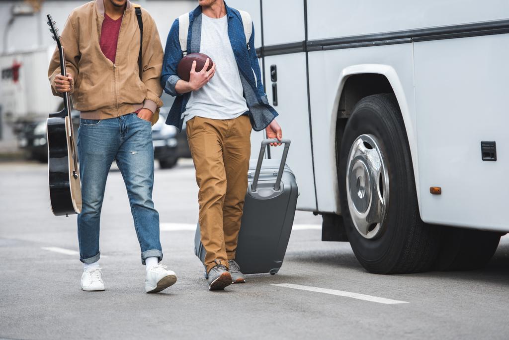 Teilansicht eines Mannes mit Rugby-Ball, der eine Rolltasche trägt, während sein männlicher Freund in der Nähe eines Reisebusses auf der Straße läuft - Foto, Bild