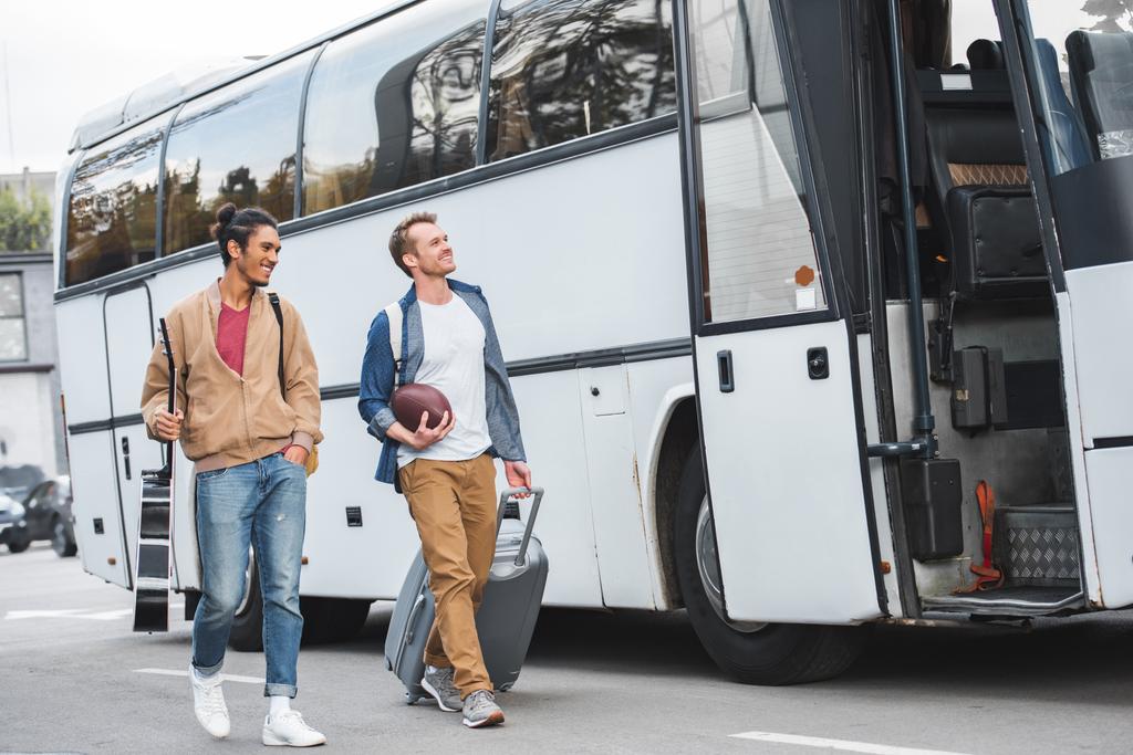 селективное внимание человека с мячом для регби, несущего сумку с колёсами, в то время как его смешанная раса мужского пола, идущего рядом с автобусом на улице
 - Фото, изображение