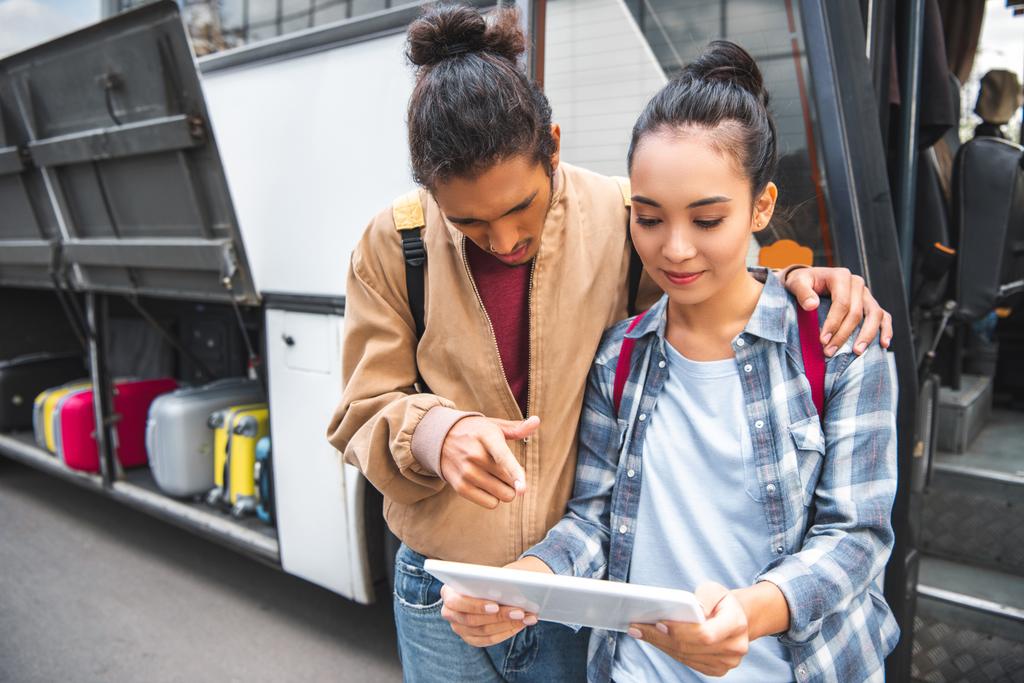 многорасовый молодой человек указывая пальцем на цифровой планшет азиатской подруге возле автобуса на улице
 - Фото, изображение